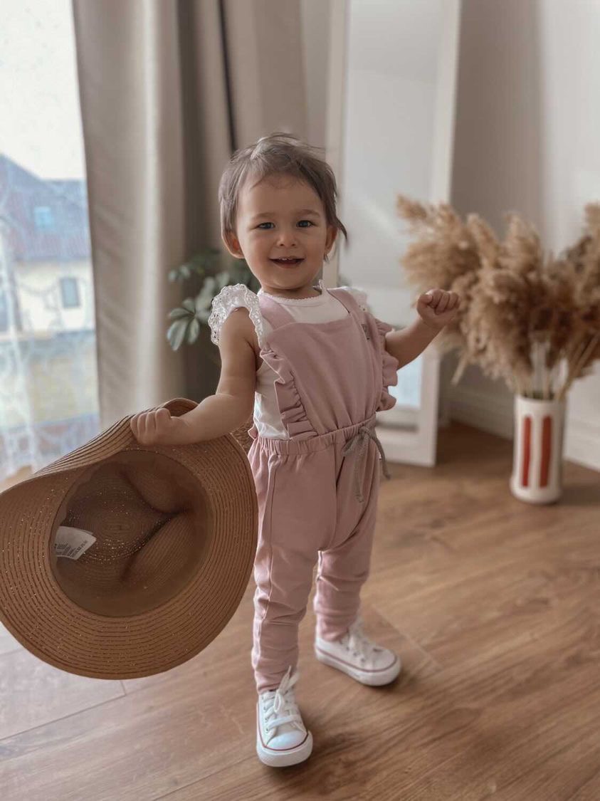 Uśmiechnięta dziewczynka w różowym kombinezonie z falbankami i białej koszulce, trzymająca słomkowy kapelusz na tle jasnego wnętrza z drewnianą podłogą i dekoracyjnym wazonem z suszonymi trawami.