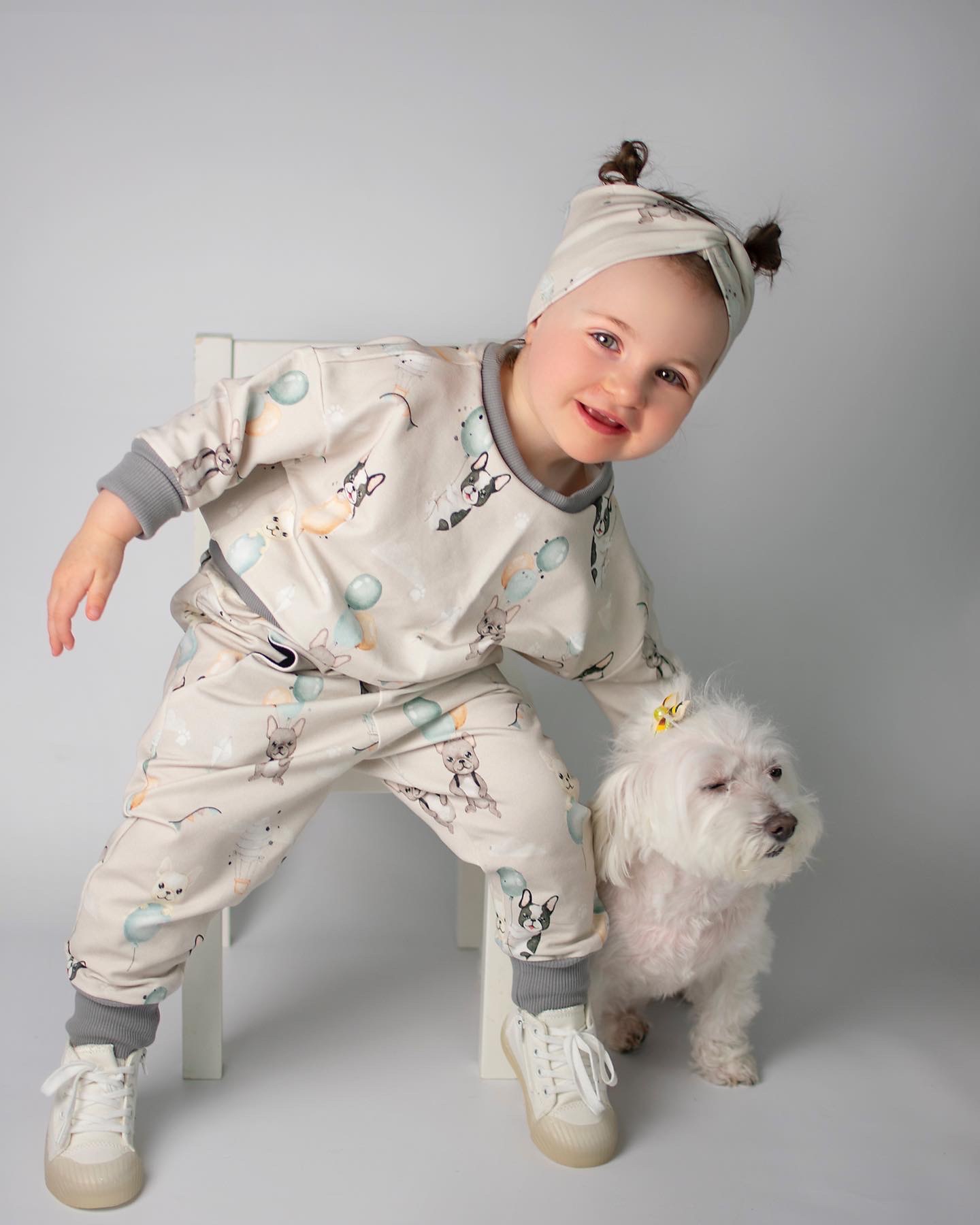 Uśmiechnięta dziewczynka w beżowym dresie z motywem buldogów francuskich i balonów, opierająca się o białe krzesło, obok niej biały pies z kokardką, na białym tle.