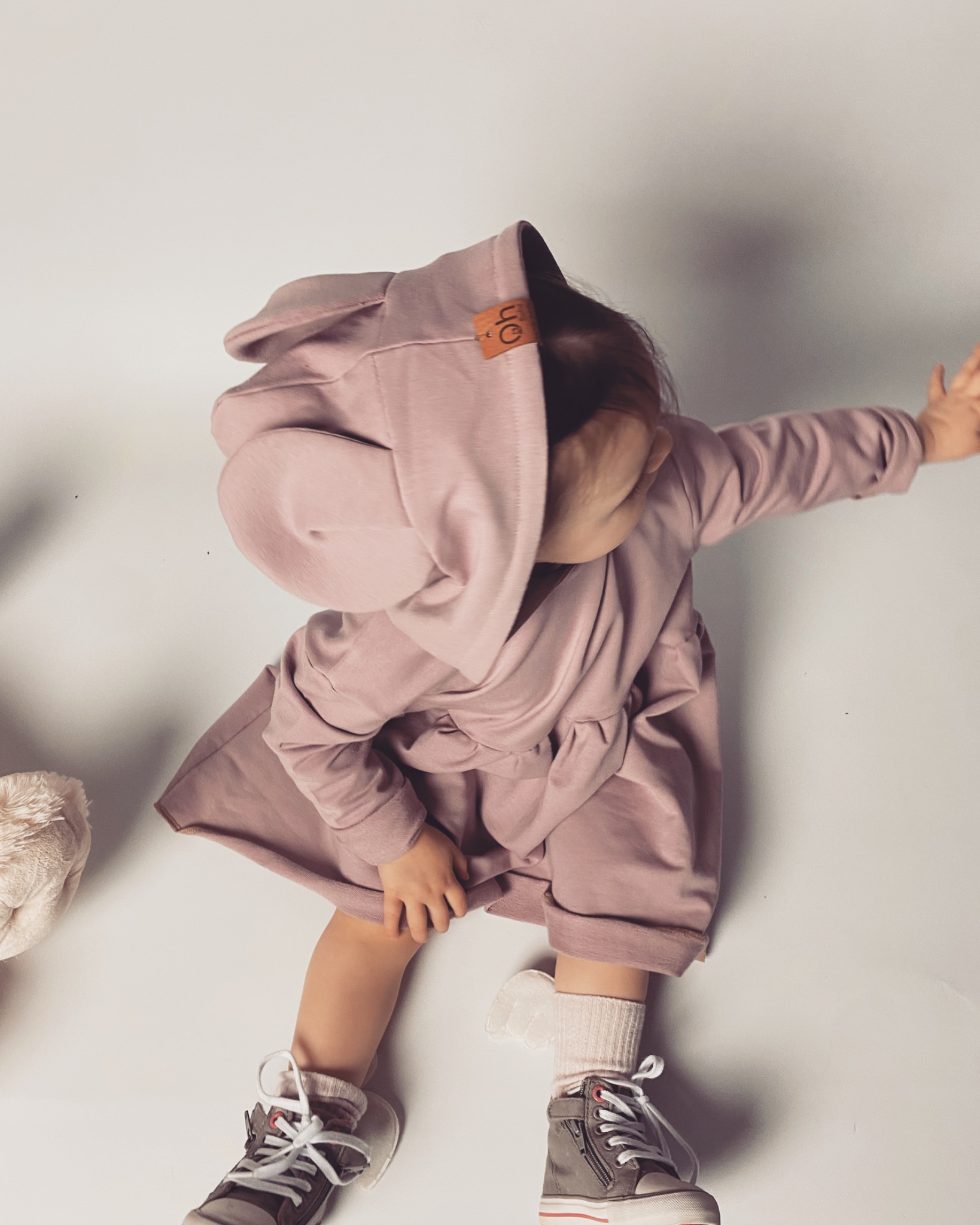Dziewczynka w pudroworóżowej sukience z kapturem i uszami, siedząca na białym tle obok pluszaka, w szarych trampkach ze sznurowadłami.