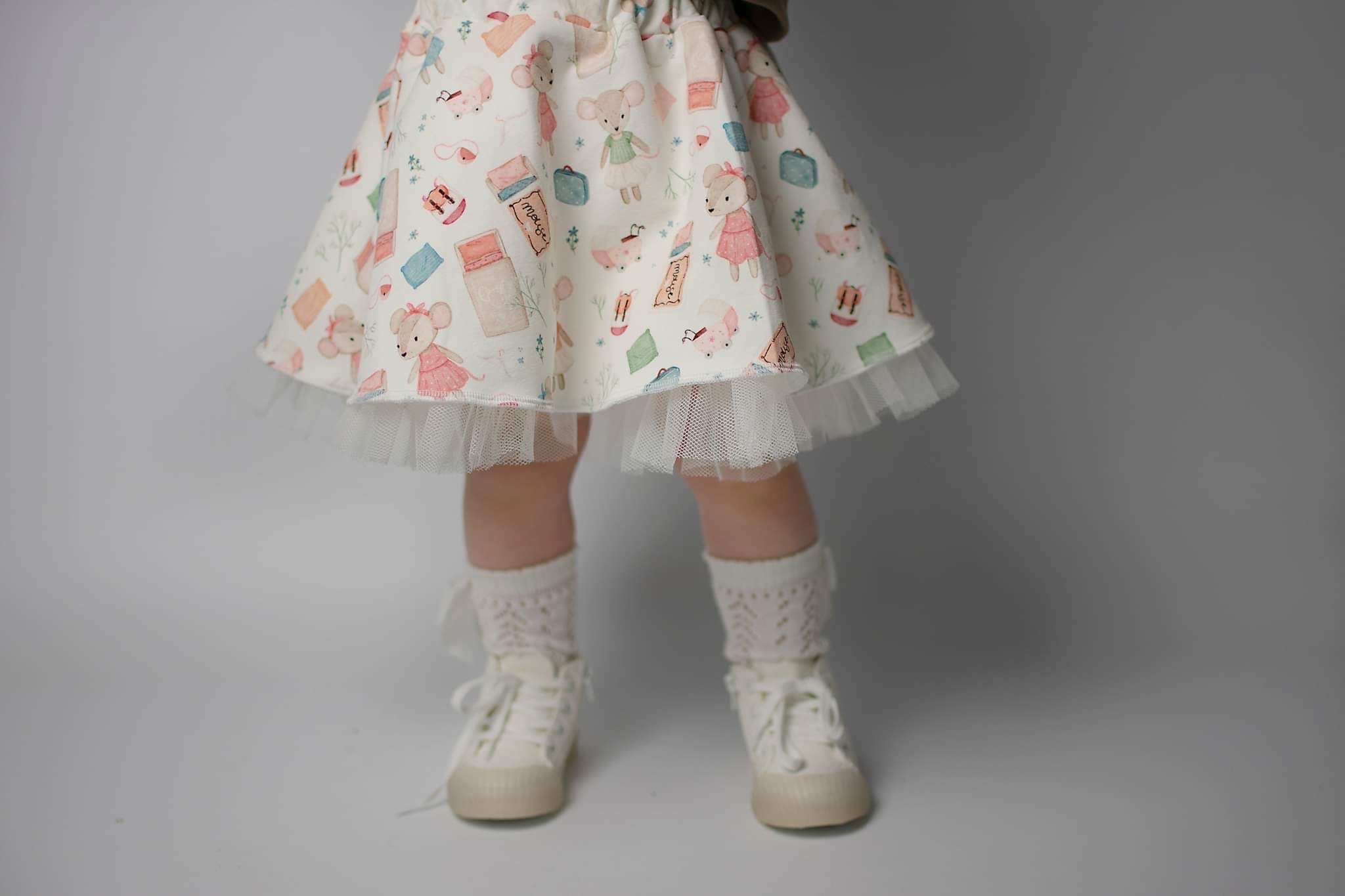 Dziewczęca spódniczka z motywem myszek i walizek, wykończona tiulem, w komplecie z ażurowymi skarpetkami i płóciennymi bucikami na jasnej podłodze.