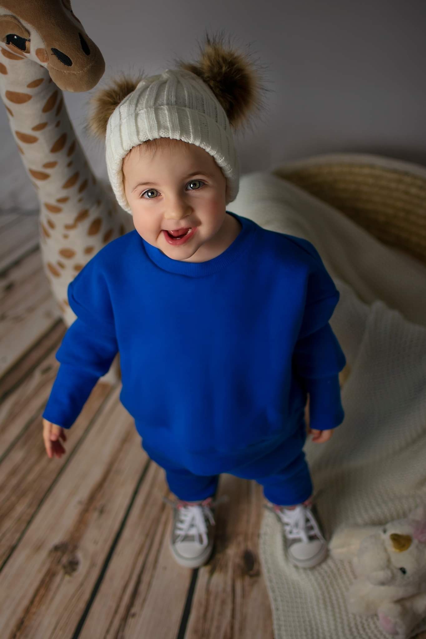 Uśmiechnięte dziecko w niebieskim dresie i białej czapce z pomponami, stojące na drewnianej podłodze obok pluszowej żyrafy.