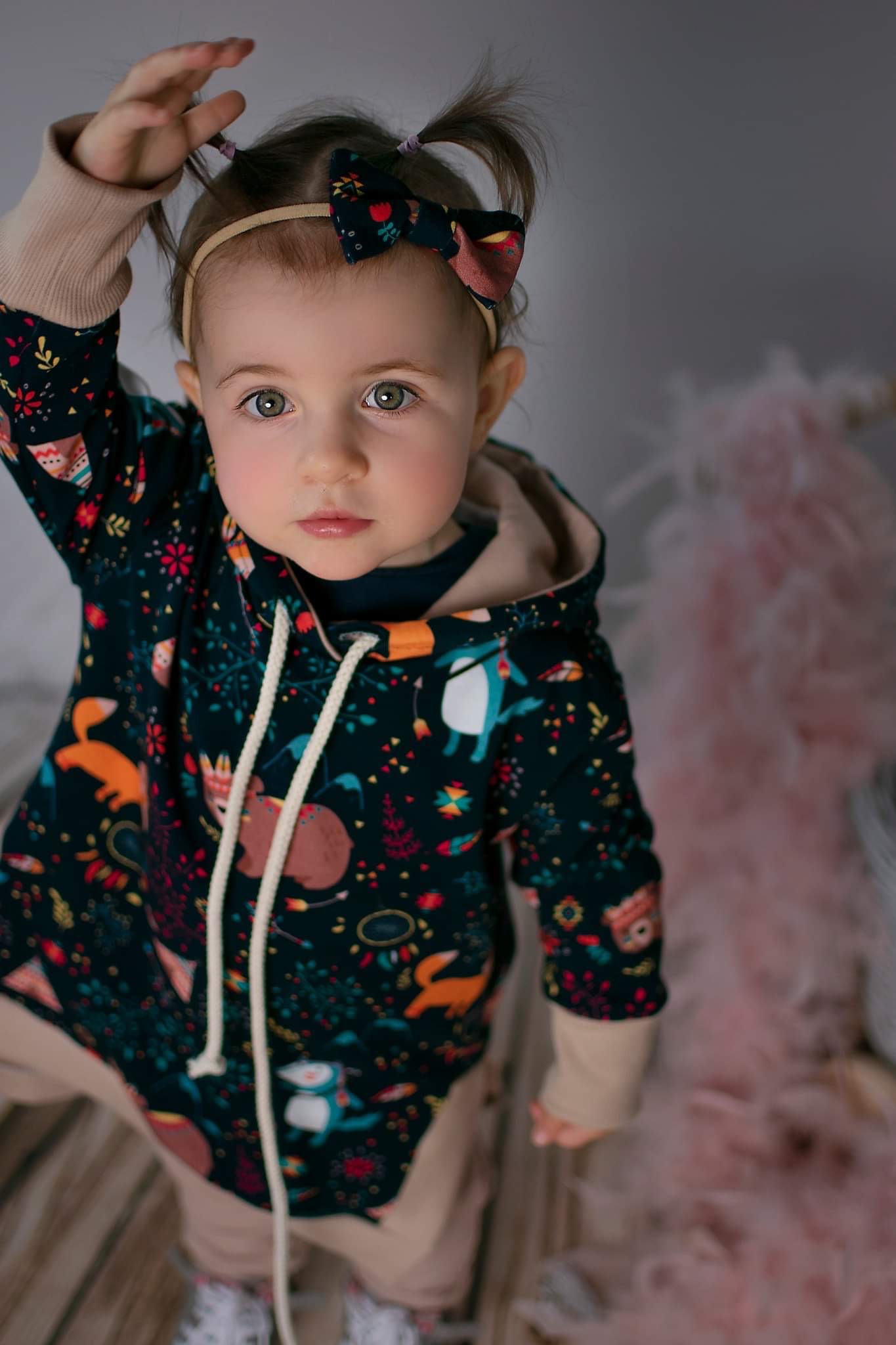 Urocza dziewczynka w ciemnej bluzie z kapturem, zdobionej wzorami zwierząt i roślin, z kokardką we włosach, patrzy w górę, unosząc rączkę.