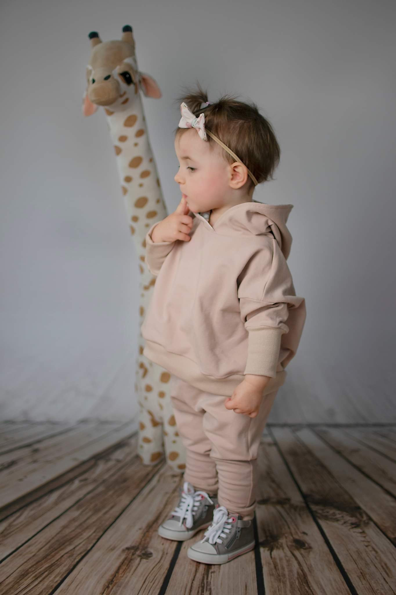 Dziewczynka w beżowym dresie z kapturem i kokardą we włosach, stojąca obok pluszowej żyrafy na drewnianej podłodze, z palcem przy ustach.