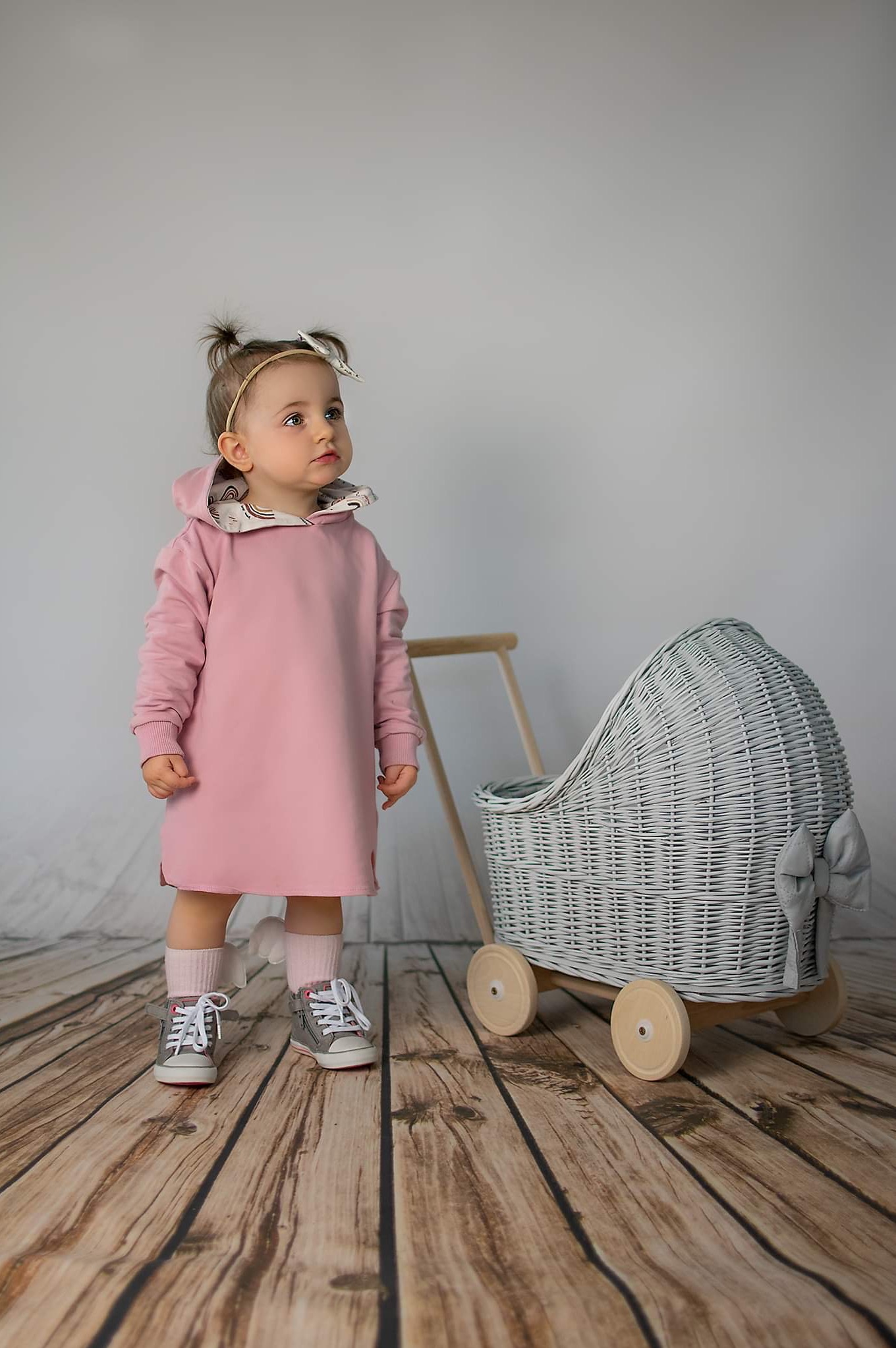 Dziewczynka w różowej sukience z kapturem, białej opasce i szarych butach stoi obok wiklinowego wózka dla lalek na drewnianej podłodze, spoglądając w górę.