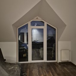 Michał Zagrabiński - Białe okno PCV w szczycie poddasza, widok z wnętrza. Podłoga drewniana, grzejnik na ścianie. Część mebli zakryta folią ochronną. Widoczne drewniane belki stropowe.