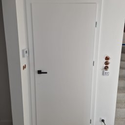 Michał Zagrabiński - Białe, minimalistyczne drzwi wewnętrzne z czarną klamką, zamontowane w odnowionym wnętrzu. Widoczne niedokończone prace elektryczne i fragment podłogi w jodełkę.