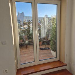 Michał Zagrabiński - Białe okno PCV z widokiem na taras z roślinami i meblami ogrodowymi. Drewniany parapet i schodek. Jasne wnętrze, słoneczny dzień.