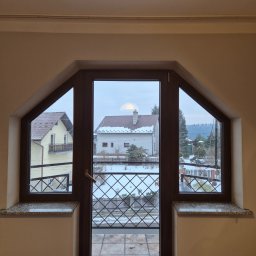 Michał Zagrabiński - Nietypowe okno PCV z balkonem i widokiem na zimowy krajobraz. Trapezoidalny kształt okna z brązową ramą i kratką dekoracyjną na balkonie.