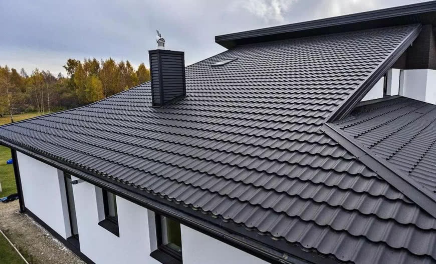 Nowoczesny dom z ciemnoszarą blachodachówką, widoczny komin z wiatrowskazem i zamontowane rynny w kolorze dachu.