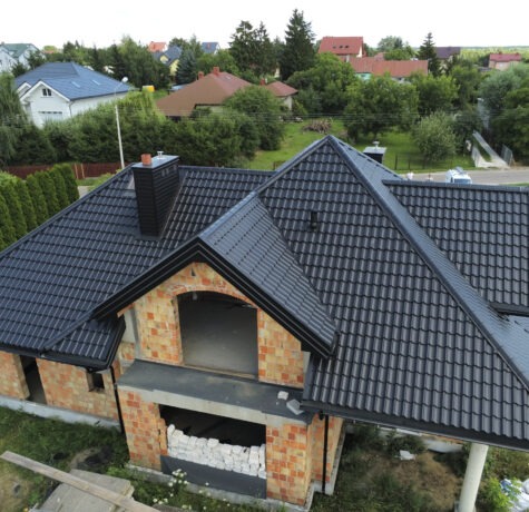 Nowy, czarny dach z dachówki ceramicznej na budynku w trakcie budowy, widoczny komin i surowa cegła, otoczenie stanowią domy jednorodzinne i zieleń.