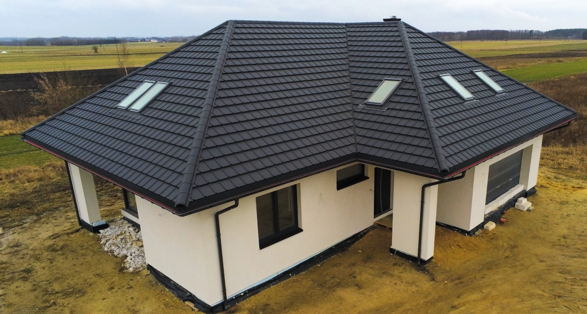 Nowoczesny dom jednorodzinny z grafitowym dachem pokrytym dachówką modułową i trzema oknami dachowymi, widok z góry na tle polnych łąk.