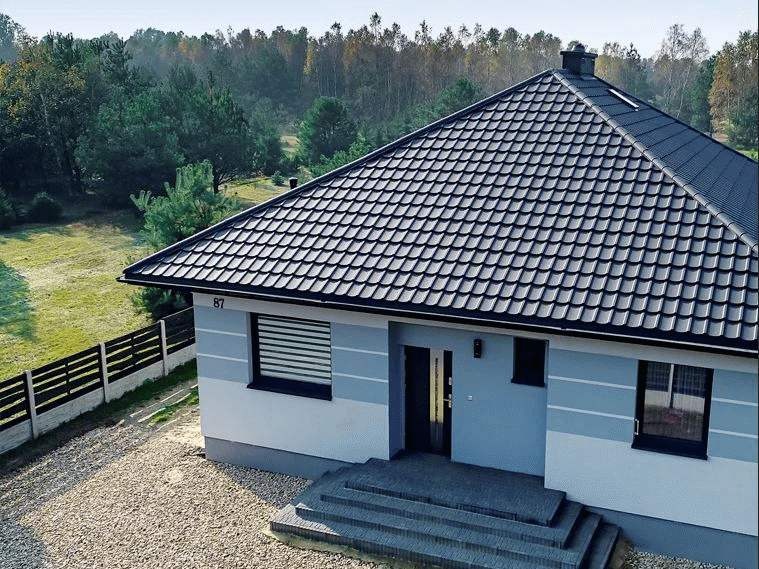 Dom jednorodzinny z ciemnoszarą dachówką, jasnoszarą elewacją z poziomymi pasami i numerem 87, otoczony zielenią i ogrodzeniem.