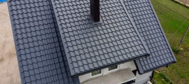 Widok z góry na dom z ciemnoszarą, metalową dachówką, kominem i rynnami, otoczony zielonym trawnikiem.