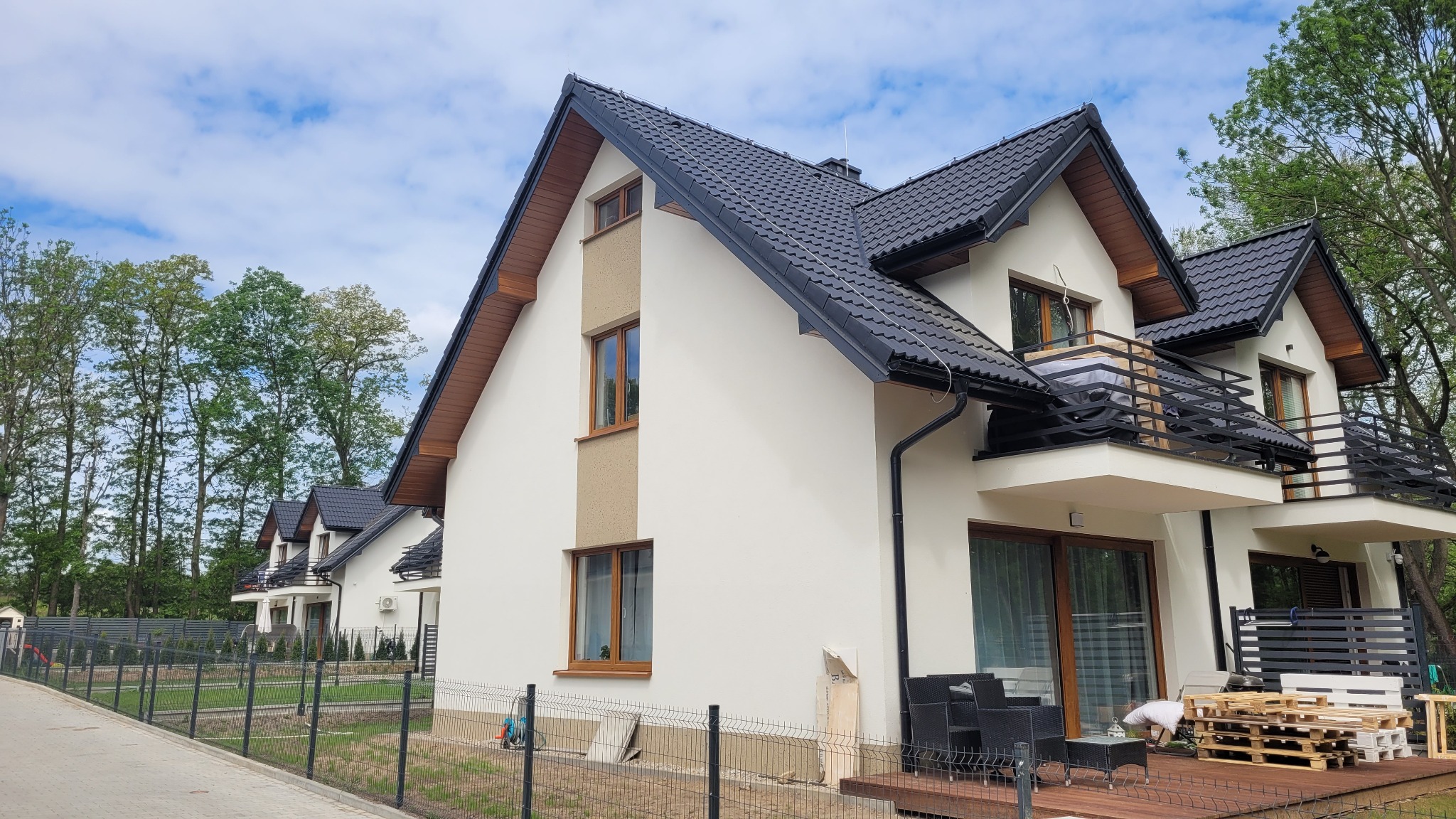 Nowoczesny dom w zabudowie szeregowej z białymi ścianami, czarnym dachem i drewnianymi oknami, widok z zewnątrz na elewację i taras z meblami ogrodowymi w trakcie wykańczania.