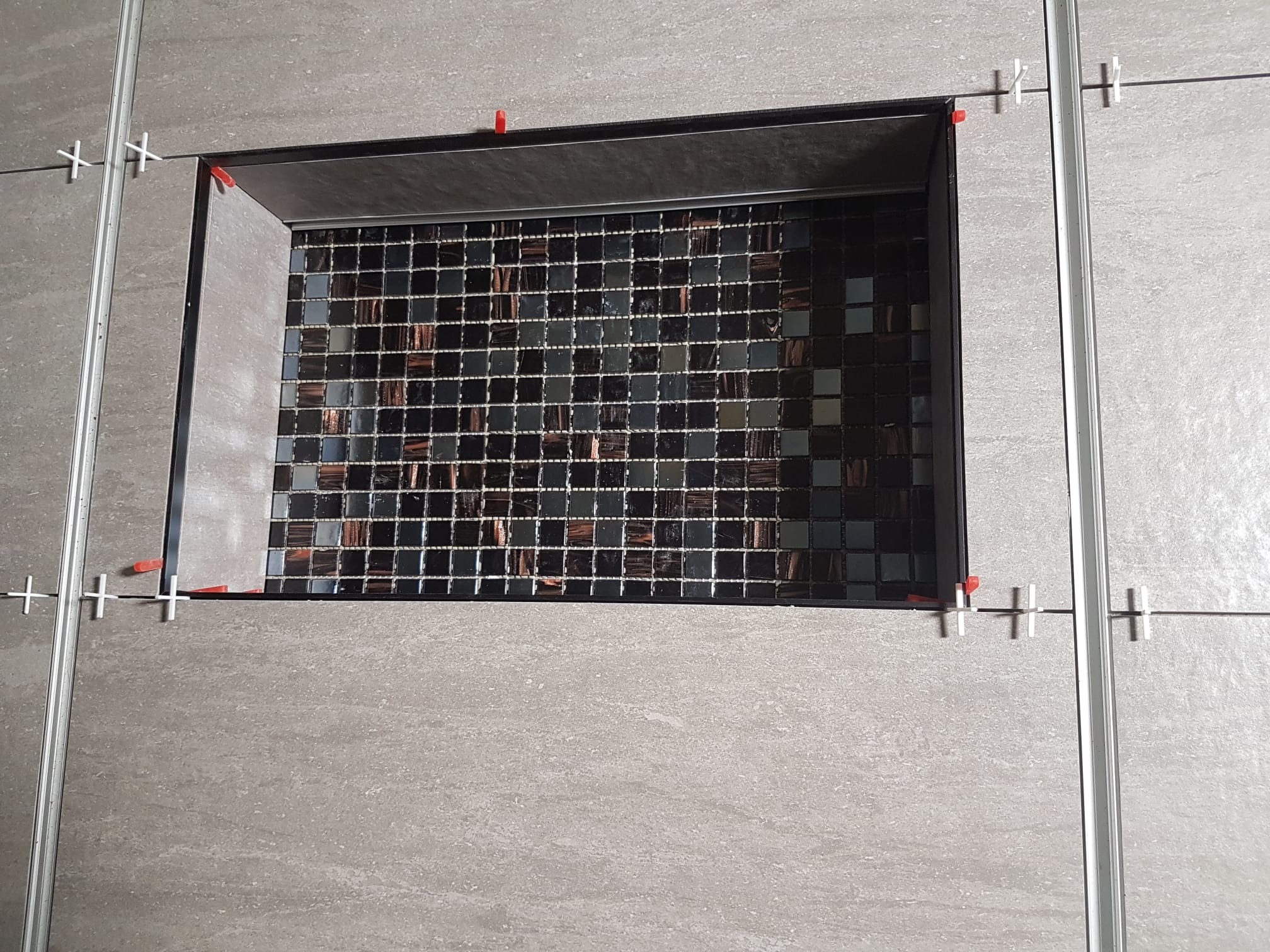 Fragment ściany z szarych płytek imitujących kamień i wnęką wyłożoną mozaiką z małych, błyszczących, ciemnych płytek. Widoczne krzyżyki dystansowe i listwy wykończeniowe.