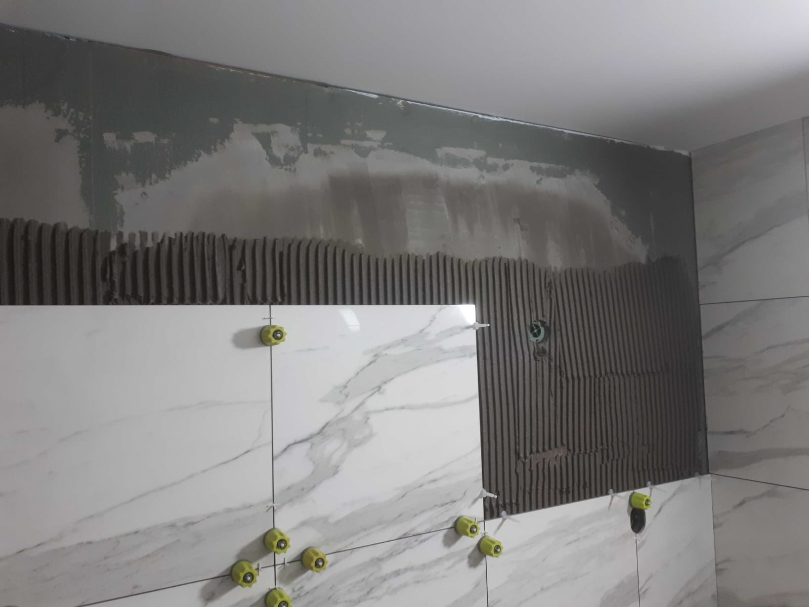 Układanie białych płytek imitujących marmur na ścianie w Częstochowie, widoczne żółte krzyżyki dystansowe i warstwa kleju.