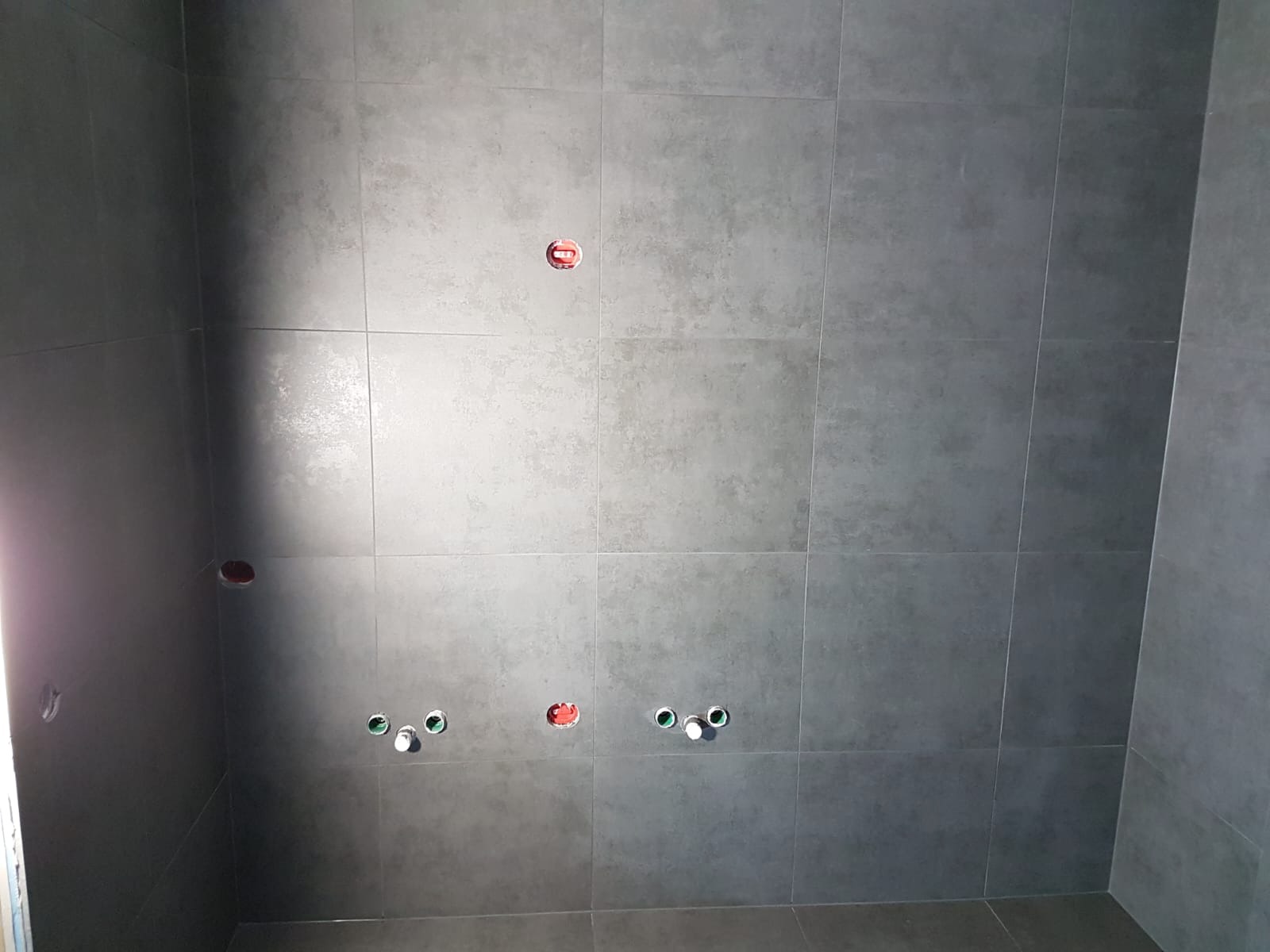 Szare, matowe płytki ceramiczne ułożone na ścianie i podłodze, widoczne otwory na instalacje wodno-kanalizacyjne i elektryczne z wystającymi rurkami i puszkami.