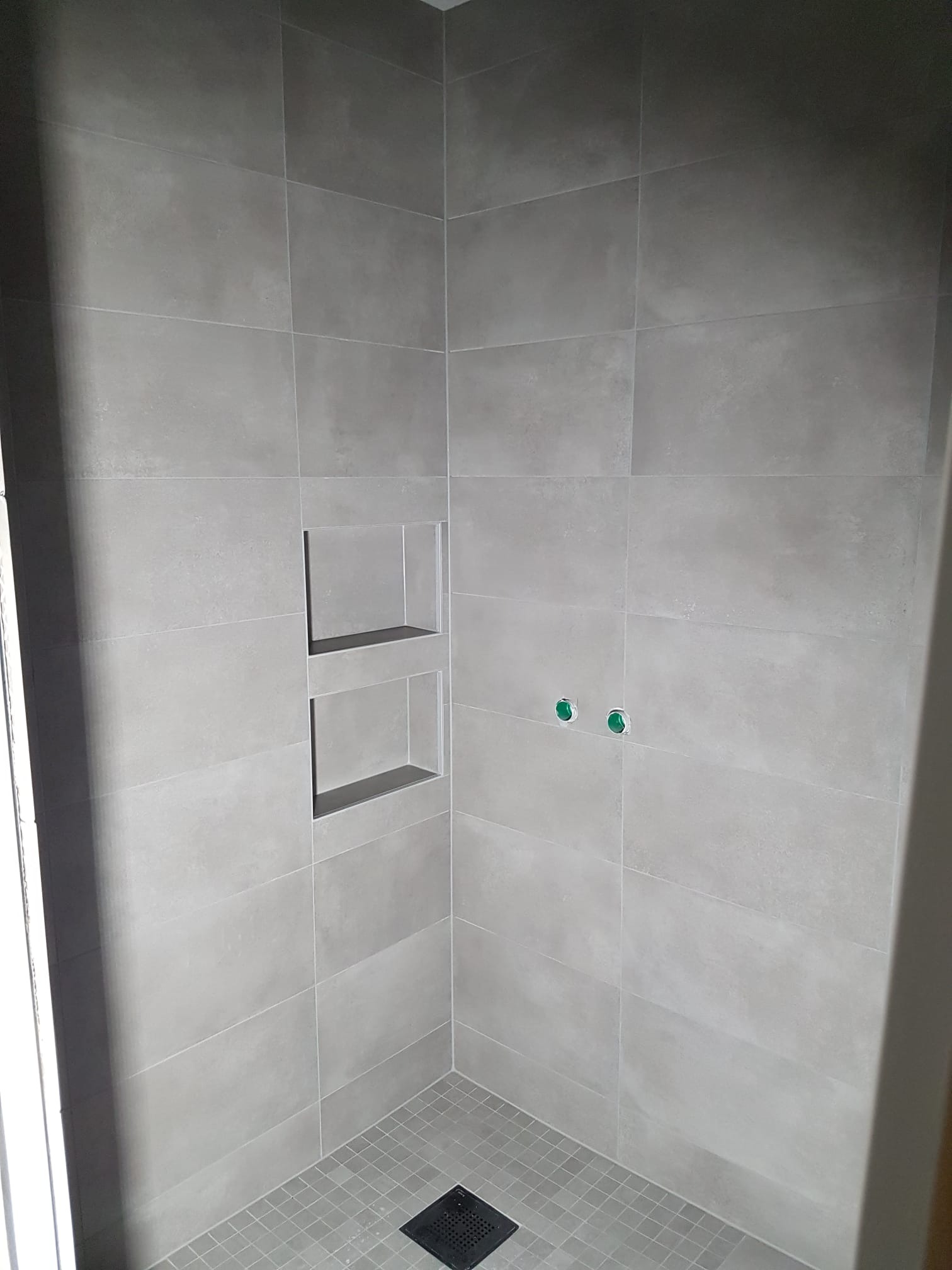 Szara kabina prysznicowa z wnękami wyłożona matowymi, kwadratowymi płytkami, odpływ liniowy w rogu, widoczne dwa zielone zaślepki na rury.