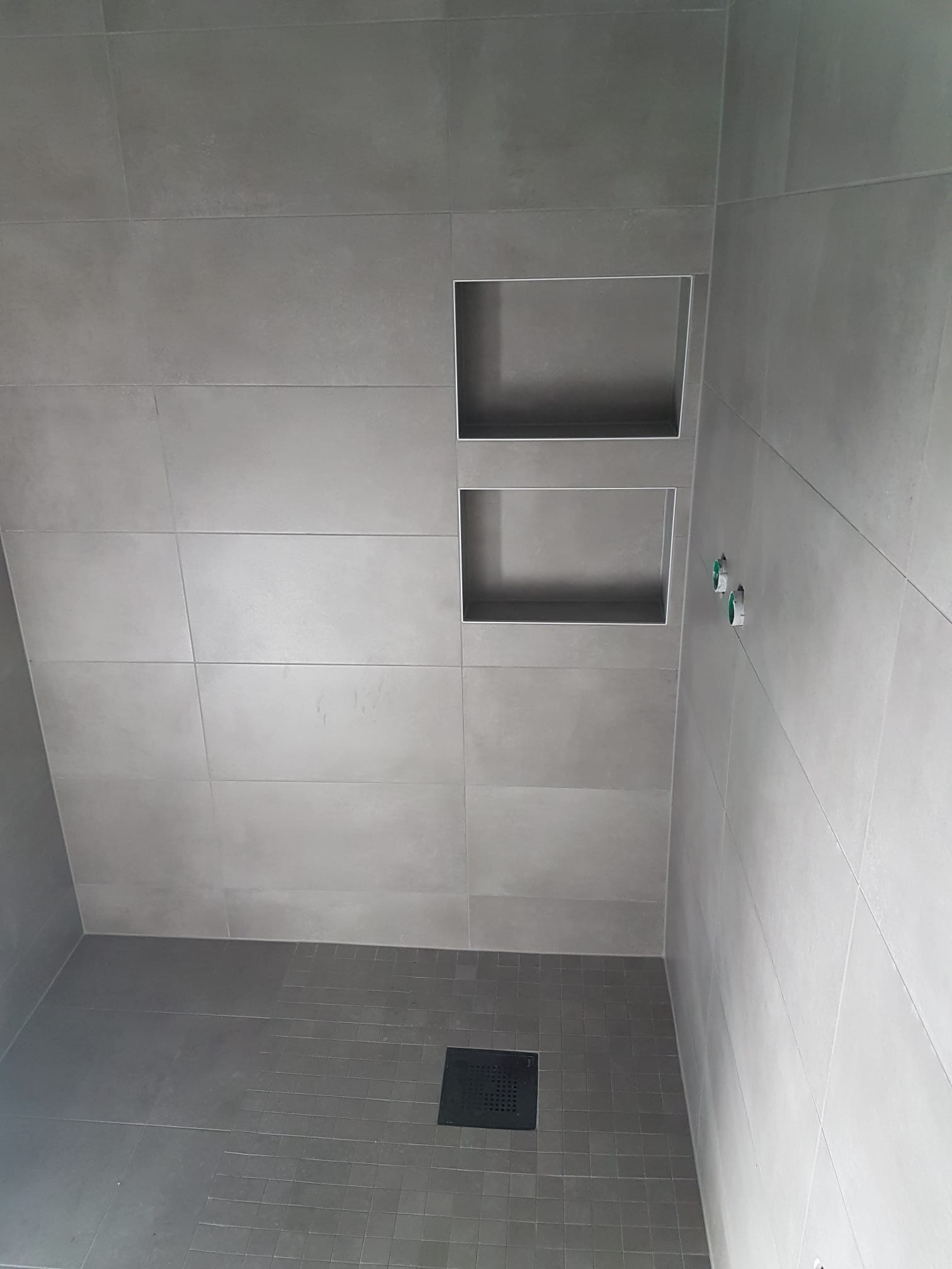 Szare, matowe płytki w kabinie prysznicowej z dwoma wnękami w ścianie i odpływem liniowym w podłodze.