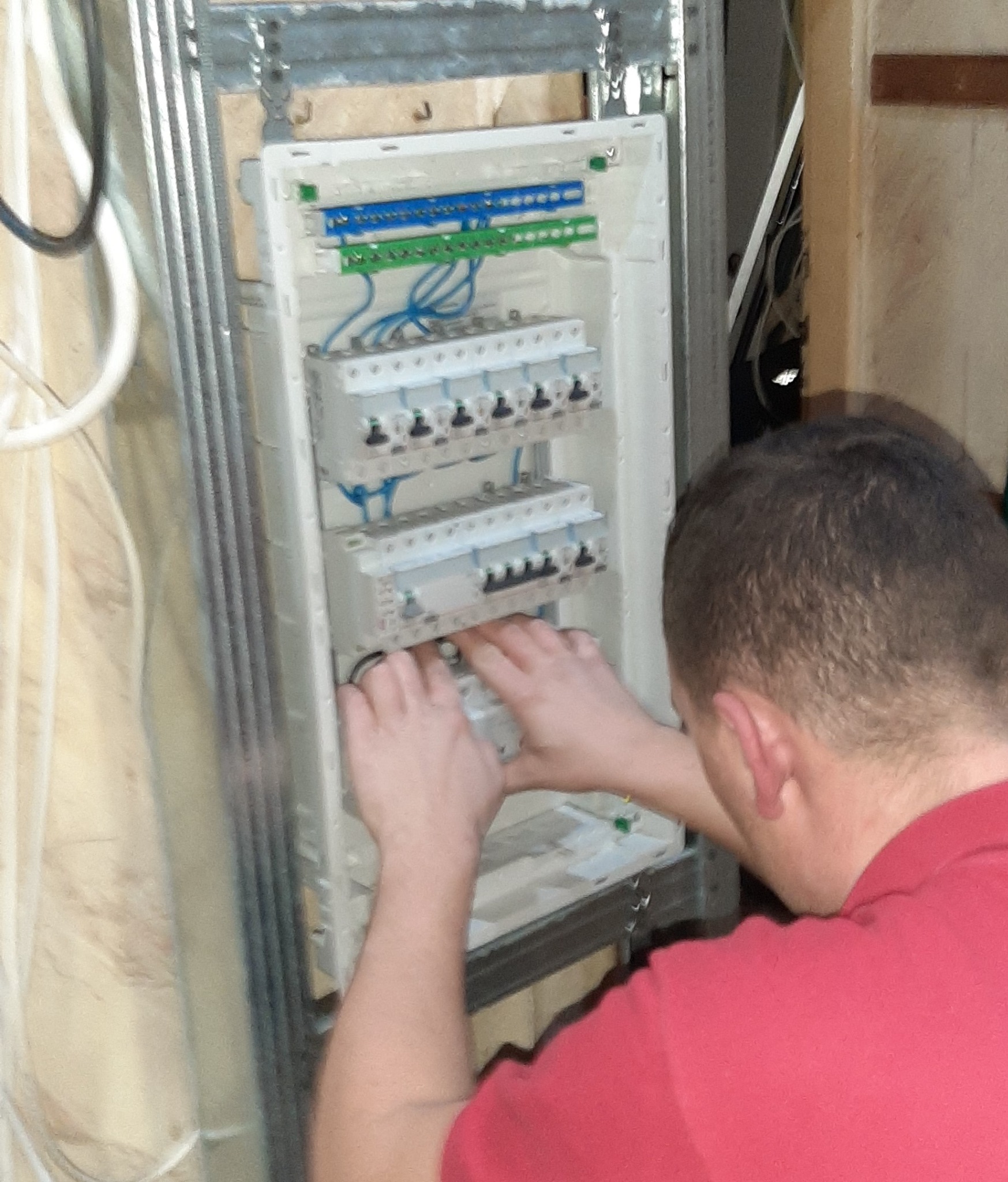 Elektryk w czerwonej koszulce instaluje zabezpieczenia w rozdzielnicy elektrycznej, widoczne przewody i listwy zaciskowe.