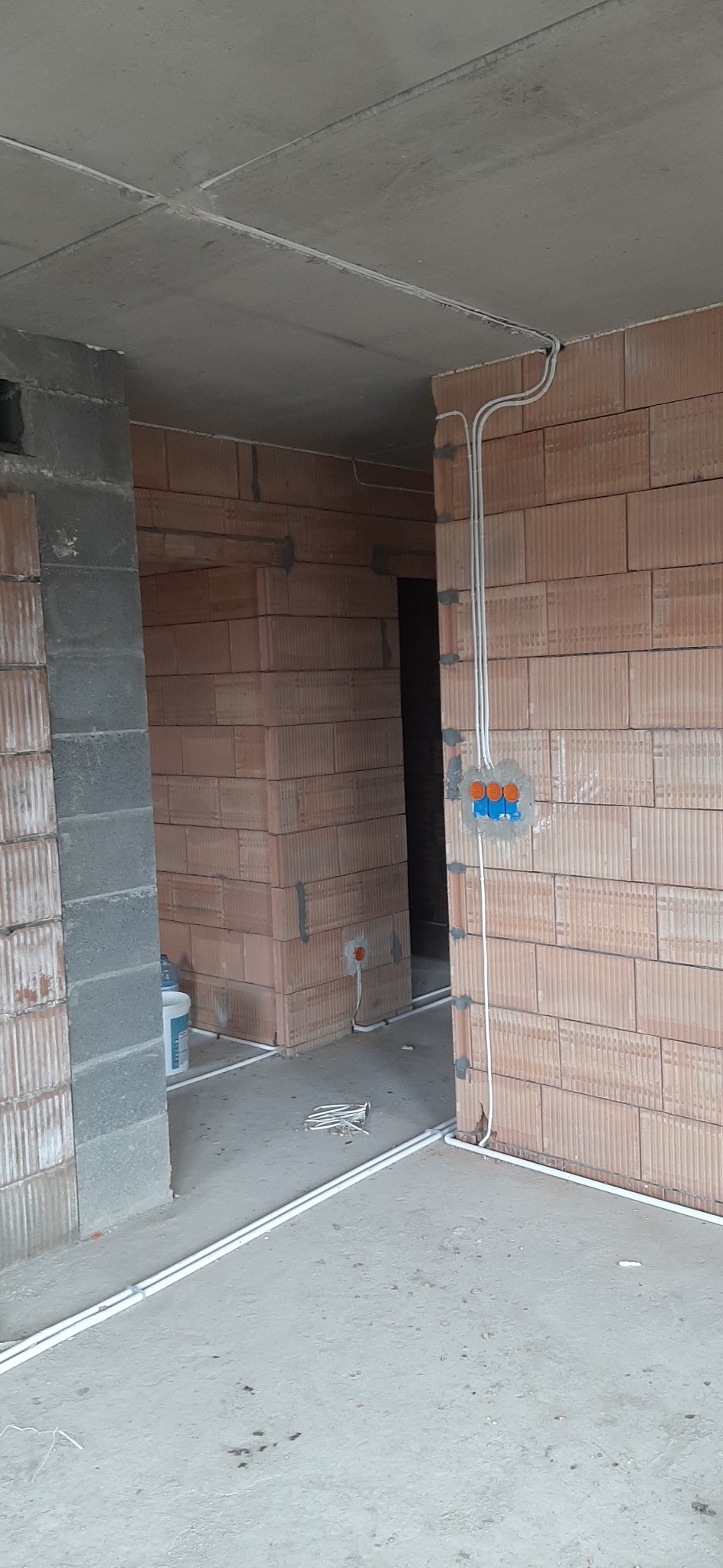 Surowy stan wykończeniowy wnętrza budynku z widocznymi przewodami elektrycznymi prowadzonymi natynkowo, puszka instalacyjna z trzema gniazdami w kolorze pomarańczowym, ściany z czerwonej cegły...