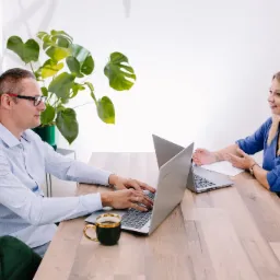 Księgowy w okularach wpisuje dane na laptopie, obok filiżanka kawy, blondynka uśmiecha się z dokumentem w dłoni, w tle rośliny doniczkowe, jasne wnętrze biura.