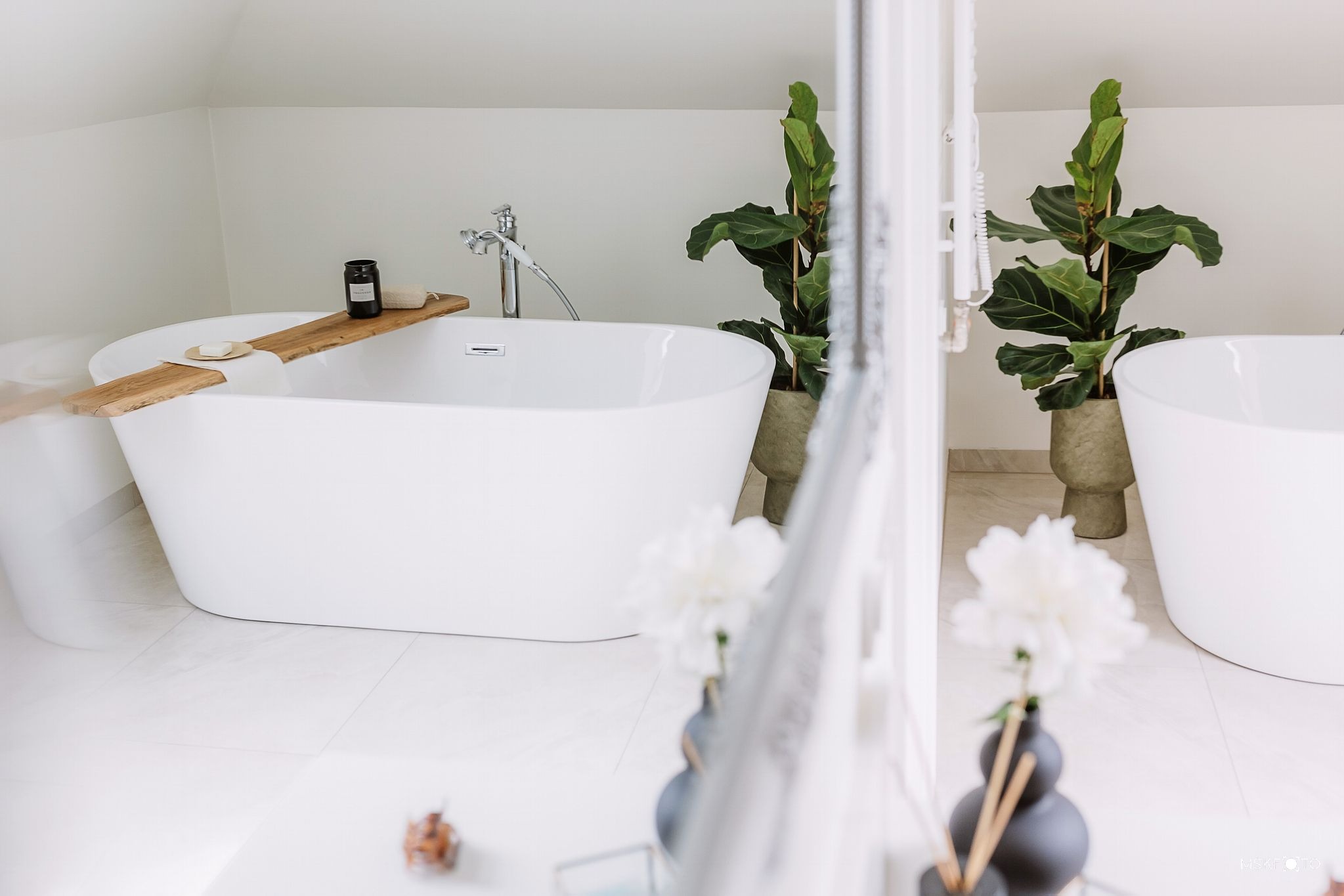 Jasna łazienka na poddaszu z wolnostojącą wanną, drewnianą półką z kosmetykami, roślinami w donicach i dekoracyjnymi kwiatami w wazonach.