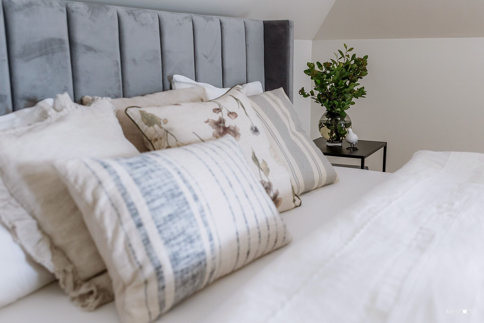 Sypialnia w neutralnych barwach z szarym zagłówkiem, dekoracyjnymi poduszkami w paski i z motywem kwiatowym oraz stolikiem nocnym z bukietem w szklanym wazonie i białą figurką ptaka.