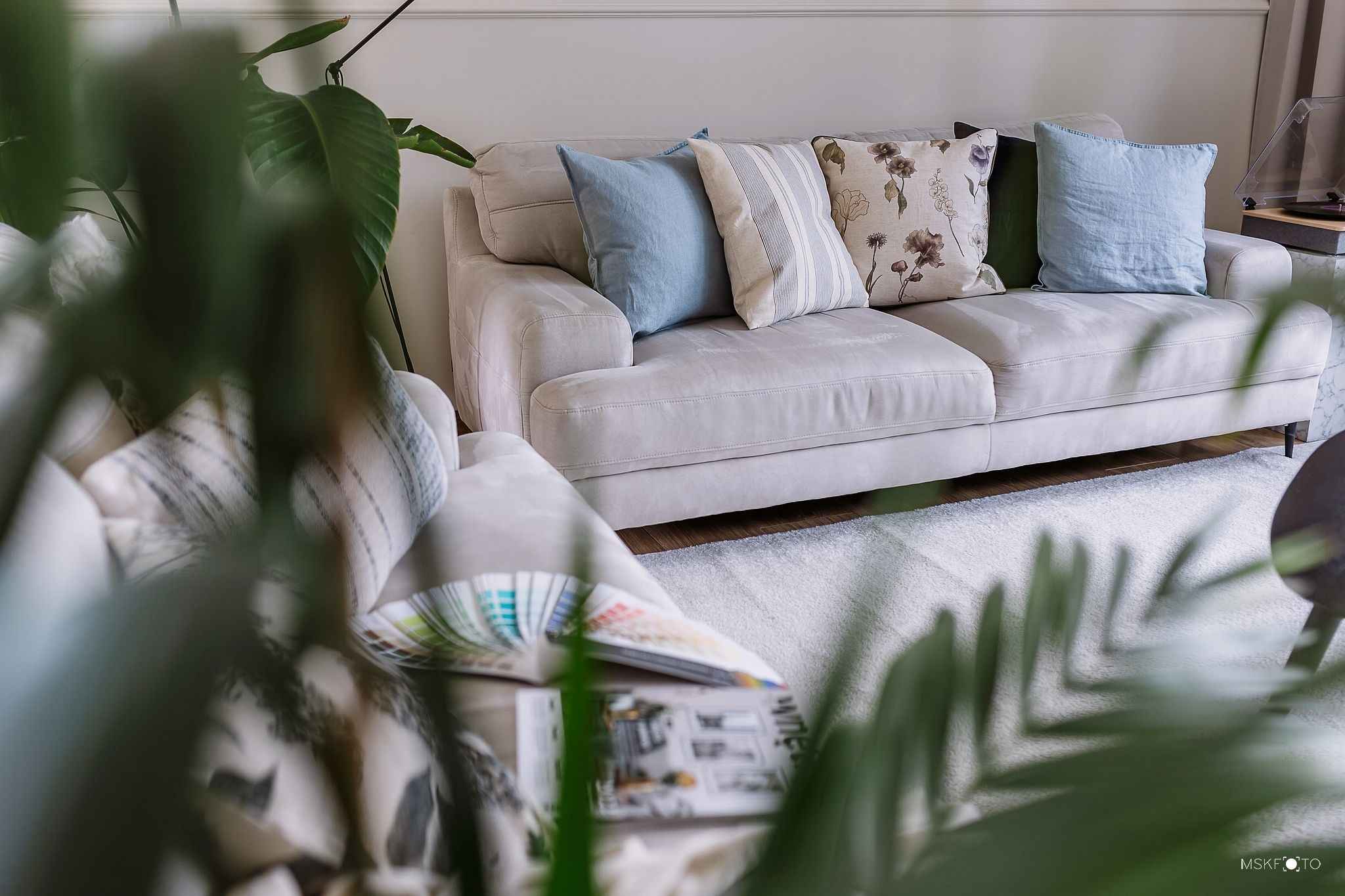 Jasnoszara sofa z poduszkami w odcieniach błękitu, beżu i brązu, na podłodze puszysty dywan, w tle gramofon, w pierwszym planie roślina doniczkowa i paleta kolorów na fotelu.