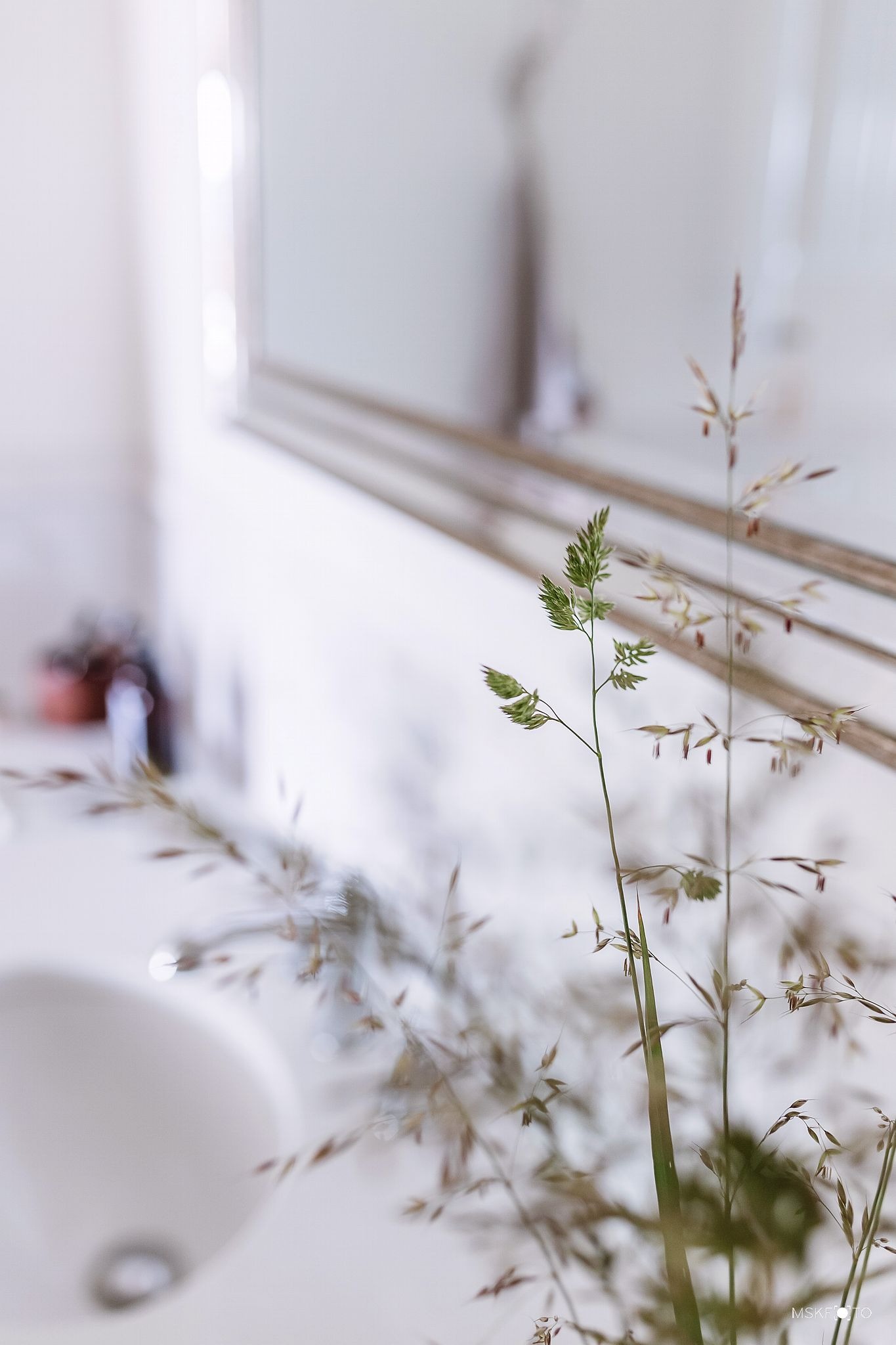 Minimalistyczna łazienka z bliska: źdźbła trawy w wazonie na tle umywalki i lustra w srebrnej ramie, miękkie światło.