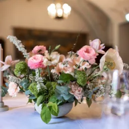 Kompozycja kwiatowa w odcieniach r&oacute;żu i bieli w okrągłym, szarym wazonie, ustawiona na białym obrusie, w tle rozmyte światło żyrandola i dekoracyjne malowidła na suficie.