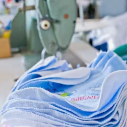 Stos niebieskich śliniaczków dziecięcych z nadrukiem leży na stole w szwalni, w tle widoczna maszyna do szycia.