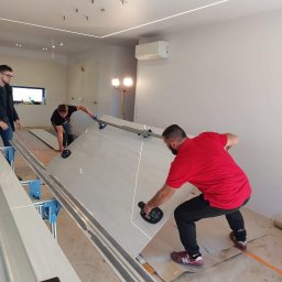 Decisio Remonty - Trzech mężczyzn przenosi płytę wielkoformatową za pomocą przyssawek w pomieszczeniu z surowym oświetleniem LED i widocznymi instalacjami elektrycznymi.