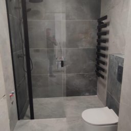 Decisio Remonty - Wykończona łazienka z szarymi płytkami imitującymi marmur, kabiną prysznicową typu walk-in z czarnymi profilami, wiszącą toaletą i grzejnikiem drabinkowym w kolorze czarnym; widoczne odbicie...