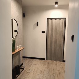 Decisio Remonty - Nowoczesny przedpokój z drewnopodobną podłogą, składanymi drzwiami, okrągłym lustrem i minimalistyczną konsolą z dekoracjami.