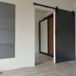 Decisio Remonty - Zamontowane przesuwne drzwi stodołowe w kolorze grafitowym, z czarnym systemem szynowym, częściowo zasłaniające wejście do garderoby z lustrzanymi drzwiami i drewnianymi panelami dekoracyjnymi...