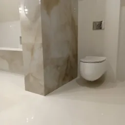 Nowoczesna łazienka z podwieszaną toaletą, obudową z płytek imitujących marmur i błyszczącymi białymi płytkami podłogowymi.