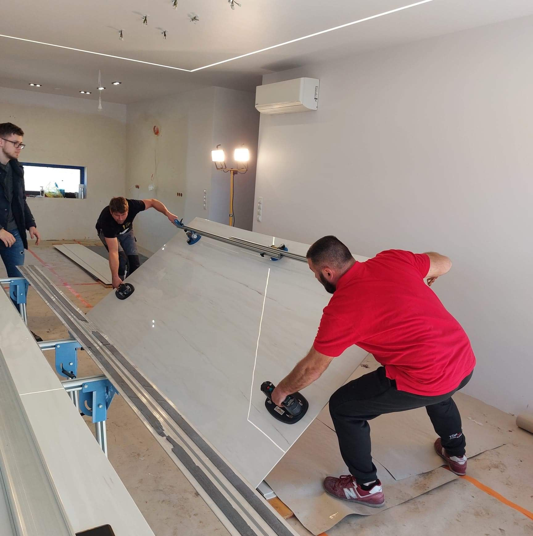 Trzech mężczyzn przenosi płytę wielkoformatową za pomocą przyssawek w pomieszczeniu z surowym oświetleniem LED i widocznymi instalacjami elektrycznymi.