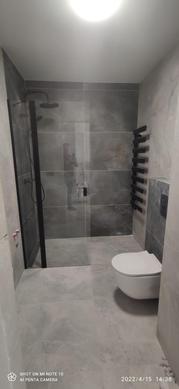 Wykończona łazienka z szarymi płytkami imitującymi marmur, kabiną prysznicową typu walk-in z czarnymi profilami, wiszącą toaletą i grzejnikiem drabinkowym w kolorze czarnym; widoczne odbicie...