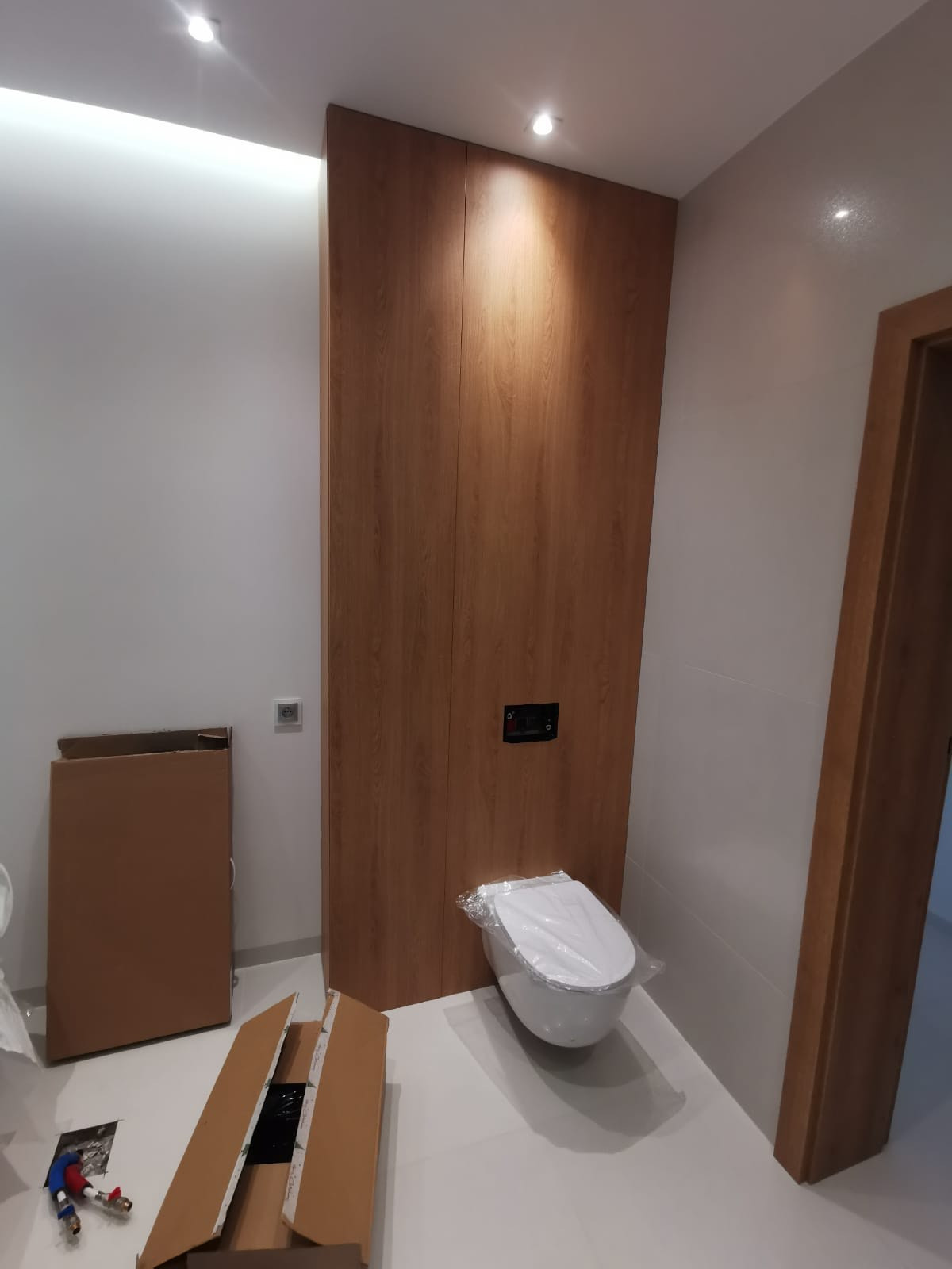 Montaż toalety w łazience: widoczna zabudowa z drewnopodobnej płyty, podtynkowa spłuczka, częściowo rozpakowana miska ustępowa z folią ochronną, kartony na podłodze.