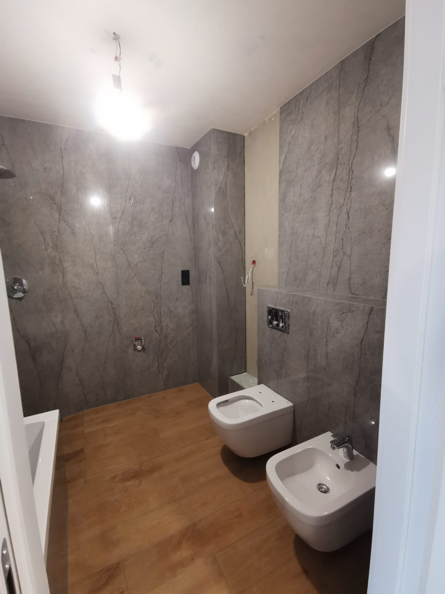 Nowoczesna łazienka w trakcie wykańczania: szare marmurowe płytki ścienne, drewnopodobna podłoga, biała umywalka i toaleta podwieszana, widoczne niedokończone prace elektryczne na suficie.