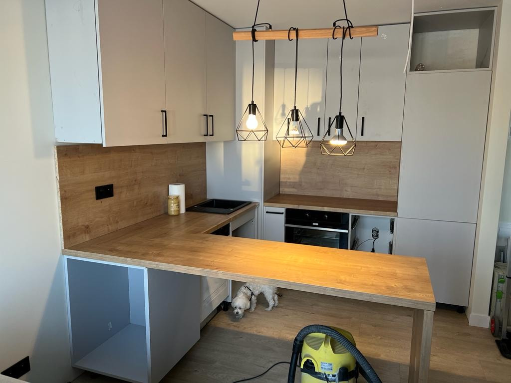 Jasna, nowoczesna kuchnia z drewnianym blatem i wiszącymi lampami; mały pies i odkurzacz w tle.