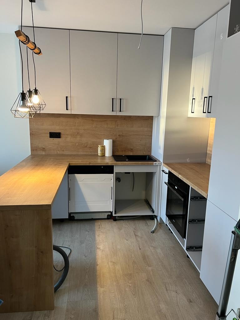 Montaż nowoczesnej kuchni z białymi szafkami i drewnianym blatem, widoczna niedokończona instalacja zlewu i otwarta zmywarka, minimalistyczne lampy wiszące.