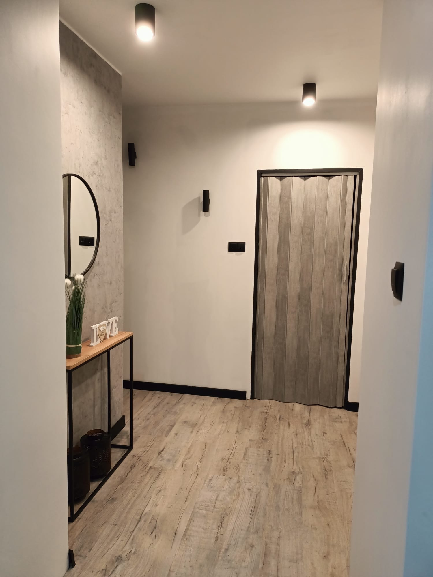 Nowoczesny przedpokój z drewnopodobną podłogą, składanymi drzwiami, okrągłym lustrem i minimalistyczną konsolą z dekoracjami.