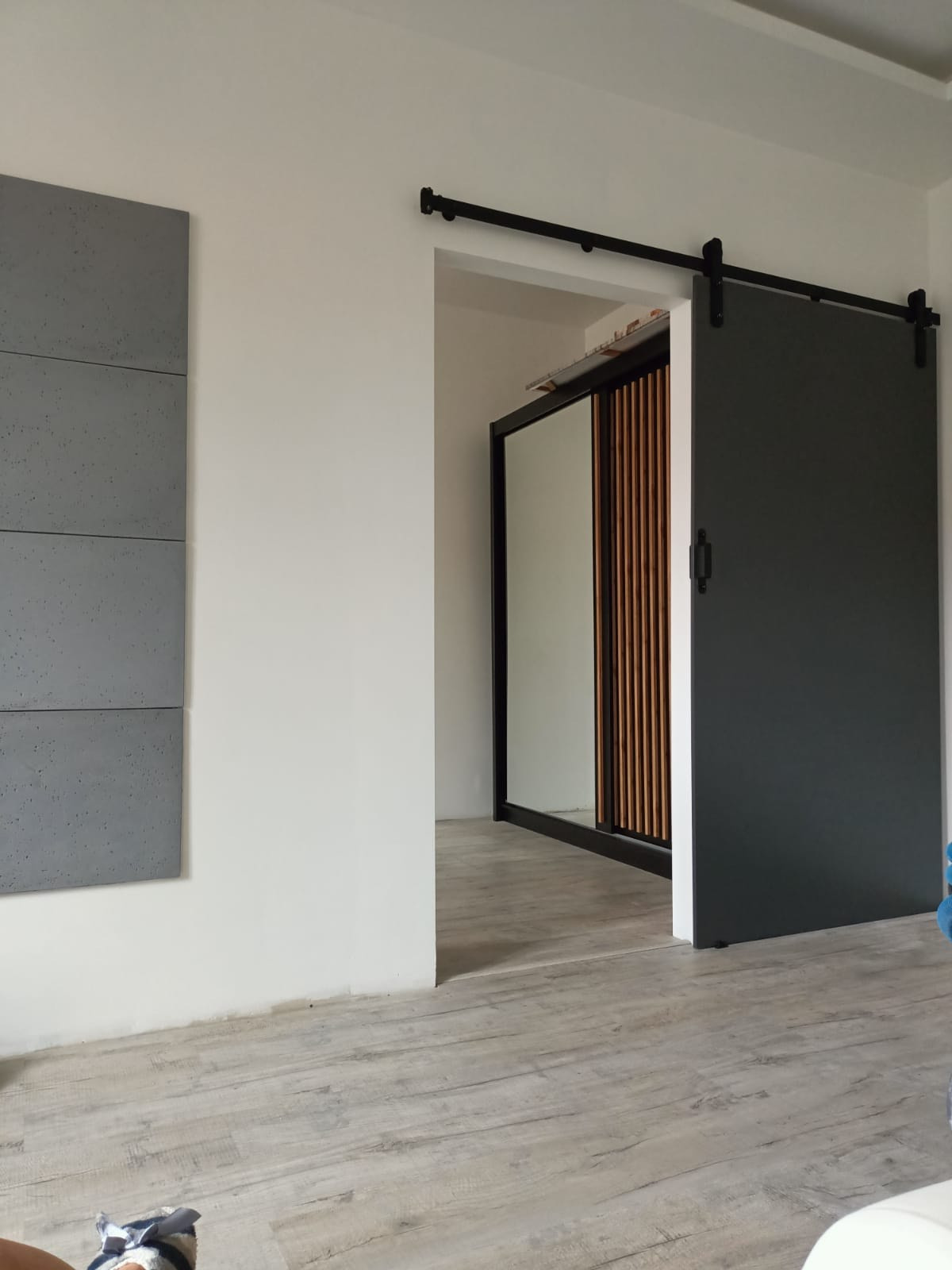 Zamontowane przesuwne drzwi stodołowe w kolorze grafitowym, z czarnym systemem szynowym, częściowo zasłaniające wejście do garderoby z lustrzanymi drzwiami i drewnianymi panelami dekoracyjnymi...