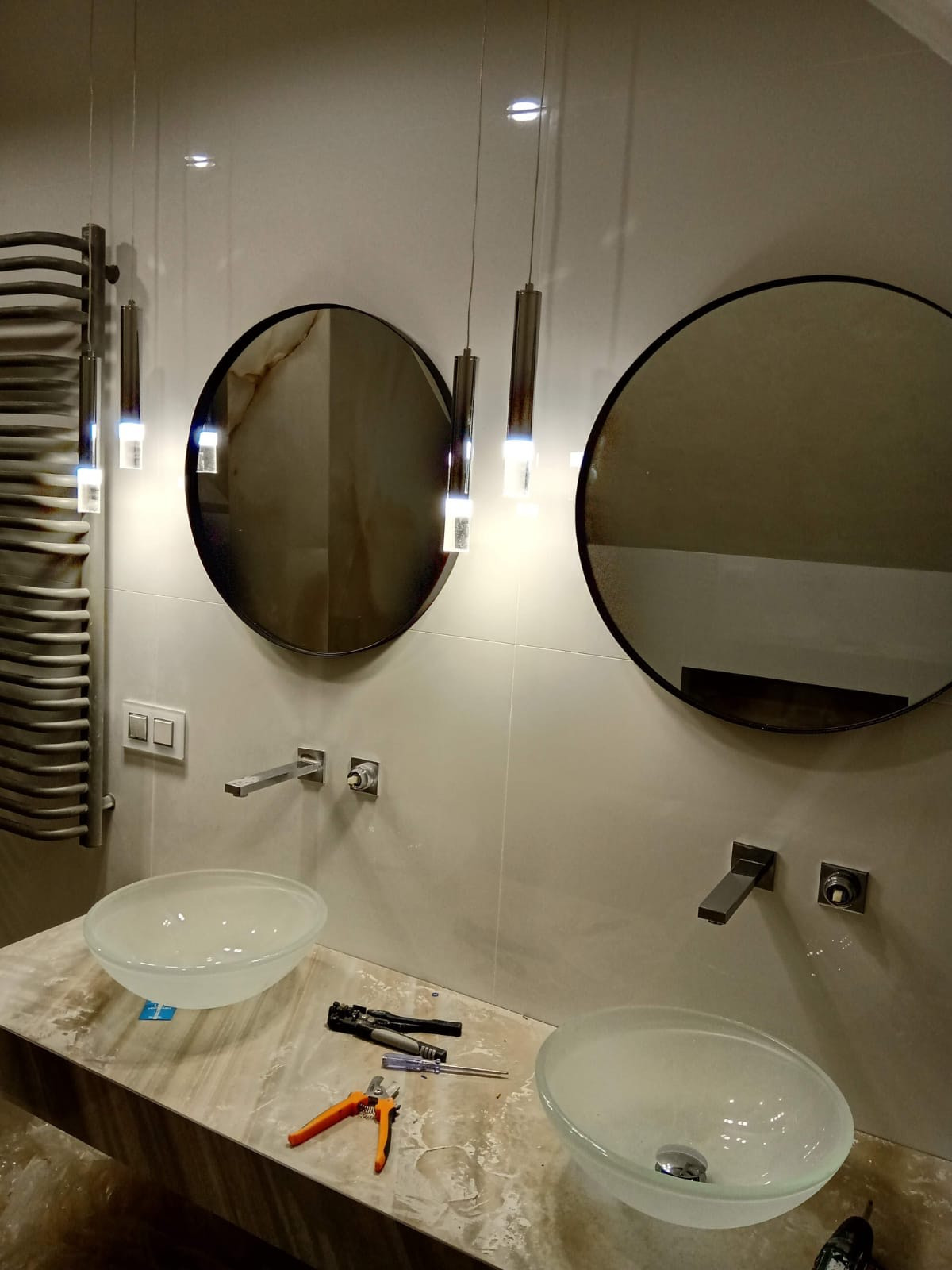 Nowoczesna łazienka w trakcie montażu: dwie szklane umywalki nablatowe, okrągłe lustra w czarnych ramach, minimalistyczne baterie ścienne, wiszące lampy tuby, grzejnik drabinkowy i narzędzia...