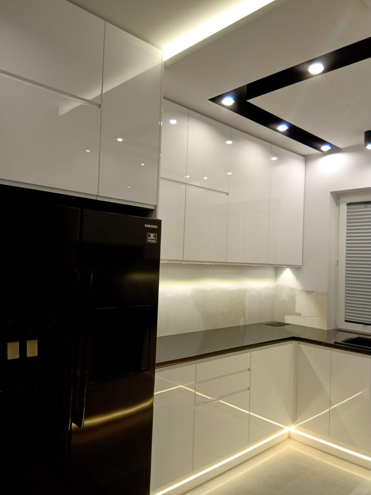 Nowoczesna kuchnia z białymi, lakierowanymi szafkami, czarnym blatem i lodówką Samsung, podświetlona listwami LED, z dekoracyjnym oświetleniem sufitowym.