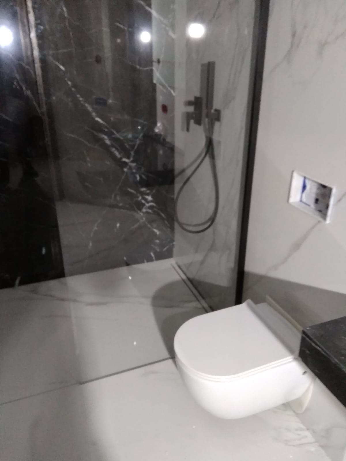 Nowoczesna łazienka z kabiną prysznicową typu walk-in wykończoną czarno-białym marmurem, wisząca toaleta i brakująca płytka wokół puszki elektrycznej.