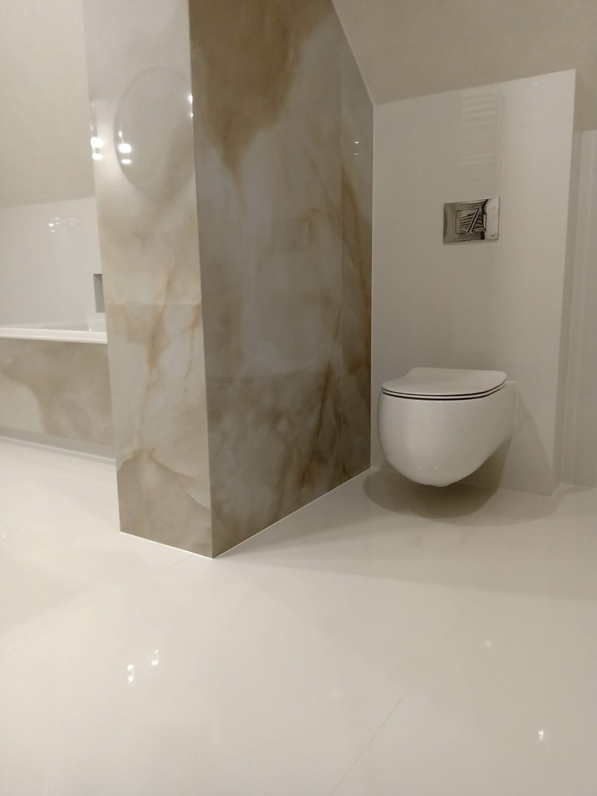 Nowoczesna łazienka z podwieszaną toaletą, obudową z płytek imitujących marmur i błyszczącymi białymi płytkami podłogowymi.