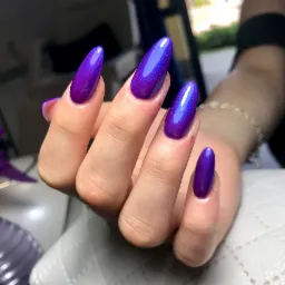 Dłoń z długimi, migdałowymi paznokciami pomalowanymi na fioletowo-niebieski, metaliczny lakier z efektem 'kociego oka', na tle jasnej, pikowanej powierzchni.